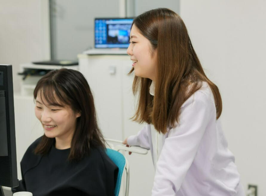 Two female accountants in Japan - one is sitting in front of a computer and the other is standing behind her while both are smiling and having a conversation